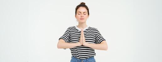 Portrait of young mindful woman meditating, holds hands clasped together, namaste gesture, practice yoga, standing over white background photo