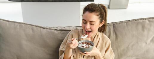 Portrait of gorgeous brunette woman sits on sofa, having her breakfast at home, smiling while eating cereals, holding bowl and spoon and laughing photo