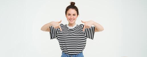Portrait of beautiful, smiling young female model, showing advertisement, pointing fingers down, standing over white background photo