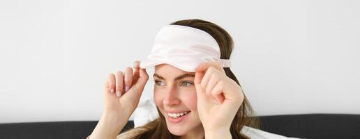 Portrait of smiling young modern woman open her eyes in morning, waking up and taking off sleeping mask, looking outside window, resting in bed, lying in bedroom photo