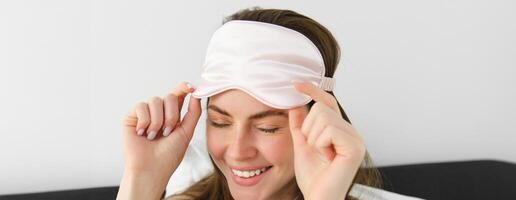 Close up portrait of gorgeous smiling woman with sleeping mask on forehead, waking up in morning, relaxing in bed, laughing and looking happy, resting on vacation in hotel room photo