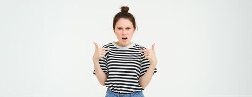 Annoyed woman pointing at herself and frowning, arguing, looking frustrated and disappointed, shouting, standing over white background photo
