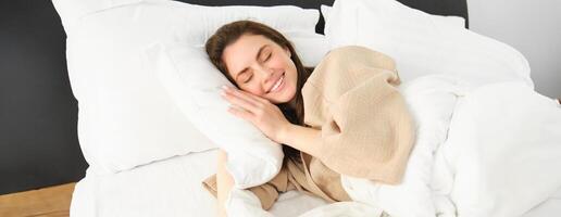Portrait of smiling brunette woman in pyjamas, sleeping in hotel bed, relaxing with pleased face, dreaming, sleeping in bedroom photo