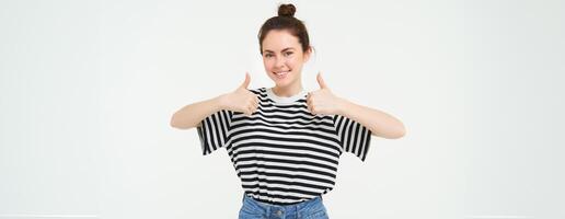 Portrait of cute girl, student shows thumbs up, recommends product, approves choice, likes item, stands over white background photo