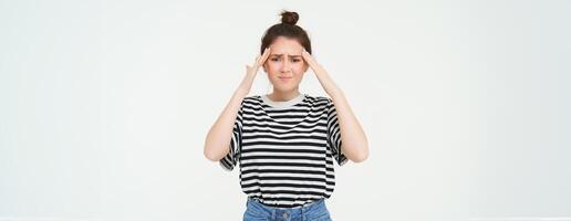 Portrait of woman with headache, touches her forehead and frowns from discomfort, stands over white background photo