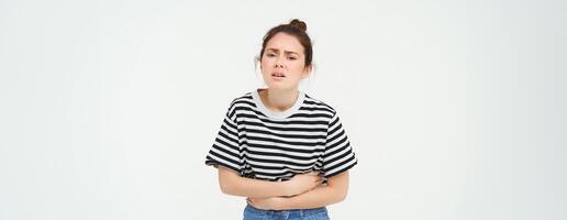 Image of young woman holding hands on her belly, feeling discomfort, menstrual period cramps, needs painkillers, stands over white background photo