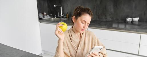 Image of attractive young woman in bathrobe, sits in kitchen, eats green apple and looks at mobile phone, uses smartphone app, orders food on application photo