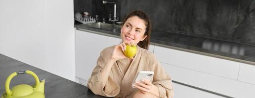 Image of attractive young woman in bathrobe, sits in kitchen, eats green apple and looks at mobile phone, uses smartphone app, orders food on application photo