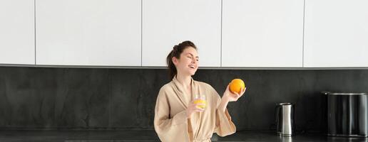 retrato de joven y sano mujer empieza su día con frutas, comiendo Fresco naranja, Bebiendo jugo en cocina, vistiendo bata de baño foto