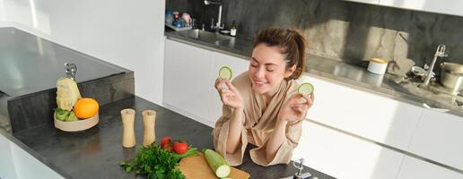 Portrait of beautiful brunette woman in the kitchen, wearing bathrobe, chopping vegetables on board, cooking healthy vegetarian food, preparing salad, making a meal photo