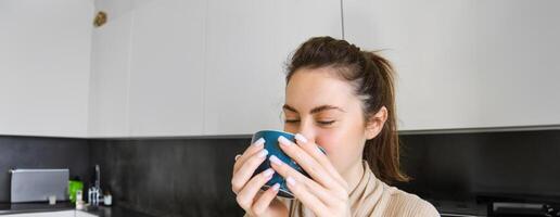 retrato de despreocupado sonriente mujer Bebiendo café, en pie en cocina con contento, satisfecho cara expresión, despertar arriba en el Mañana y teniendo capuchino foto