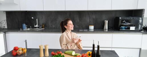 retrato de mujer en bata de baño sentado en cocina con teléfono inteligente, Cocinando cena, acecho receta en social medios de comunicación, vídeo tutorial cómo a preparar comida foto