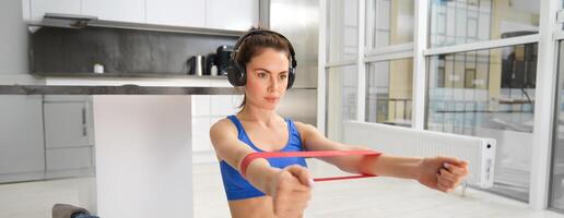 Image of hardworking sportswoman, girl doing fitness exercises at home, listening music in headphones, stretching elastic resistance band with arms, workout in living room photo