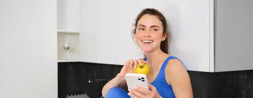 Portrait of stylish, young fitness woman, eating an apple and using mobile phone, holding smartphone, wearing sportswear photo