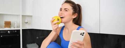 retrato de elegante, joven aptitud mujer, comiendo un manzana y utilizando móvil teléfono, participación teléfono inteligente, vistiendo ropa de deporte foto