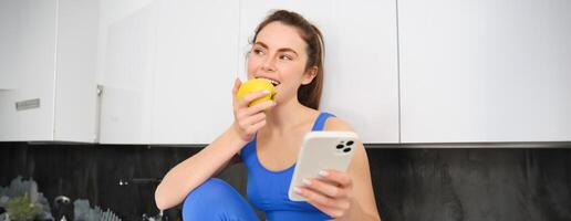retrato de saludable, deportivo joven mujer, sentado en cocina con teléfono inteligente, comiendo un manzana, mordiendo Fruta foto