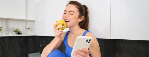 Image of stylish sportswoman, young fitness instructor sitting in kitchen and eating an apple, holding smartphone, using social media app on mobile phone photo