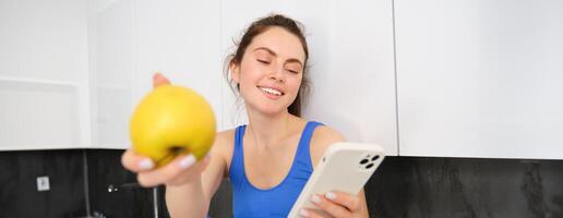 Portrait of smiling, happy fitness girl, sitting with an apple, laughing over smth on smartphone, browsing social media on mobile phone app photo