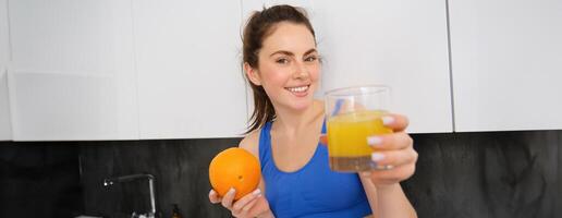 Portrait of smiling, beautiful young woman, offering glass of fresh juice, holding an orange and looking happy, fitness instructor giving you a drink photo