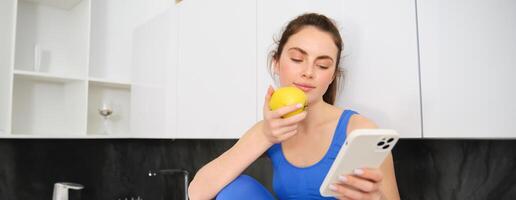 Image of stylish sportswoman, young fitness instructor sitting in kitchen and eating an apple, holding smartphone, using social media app on mobile phone photo