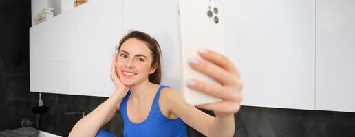 Portrait of fitness girl posing for photo, taking selfie on smartphone app, sitting in kitchen, wearing activewear photo