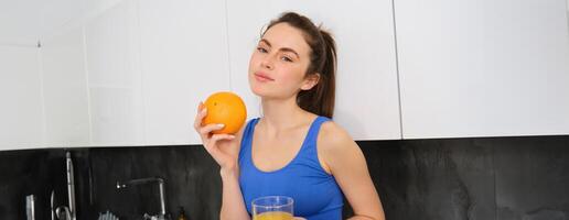 Indoor shot of young brunette woman in sportswear, drinking orange juice, holding fruit in hand, posing in kitchen photo