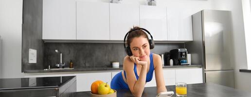 Portrait of young fitness woman with headphones, drinking orange juice in kitchen and using smartphone, listening music, getting ready for workout gym photo