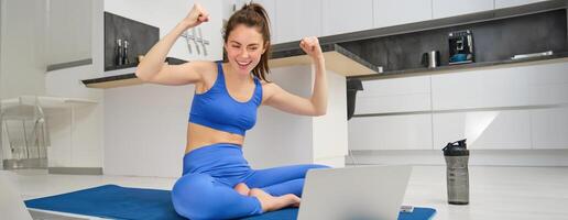 Image of excited young woman showing muscles, flexing biceps and looking at laptop screen, workout from home, fitness instructor teaching remotely, giving online yoga lesson photo