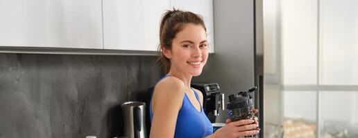 Image of young fitness instructor, woman in sportsbra and leggings, holding water bottle, drinking after workout, standing in kitchen photo