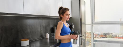 imagen de ajuste y saludable, aptitud mujer en ropa de deporte, Bebiendo agua, participación botella criba vibradora y en pie en cocina, quedarse hidratado después rutina de ejercicio foto