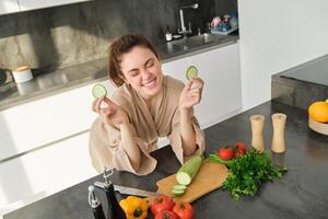 Portrait of beautiful brunette woman in the kitchen, wearing bathrobe, chopping vegetables on board, cooking healthy vegetarian food, preparing salad, making a meal photo