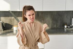Portrait of annoyed woman with knife, angry while cooking in the kitchen, frustrated while doing house chores and preparing food for family, standing in bathrobe photo