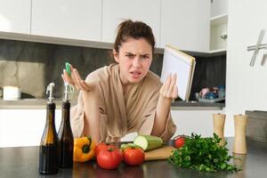 Portrait of woman cant cook, looking confused while making meal, holding recipe book, checking grocery list and staring frustrated at camera, standing near vegetables in the kitchen photo