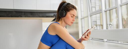 Fitness and wellbeing. Portrait of young woman doing fitness exercises, workout from home, wearing headphones and using smartphone, exercising indoors photo