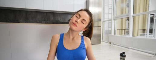 Portrait of young sportswoman, stretching her neck, warm-up before yoga exercises, doing fitness workout on rubber mat, wearing blue sportsbra and leggings photo