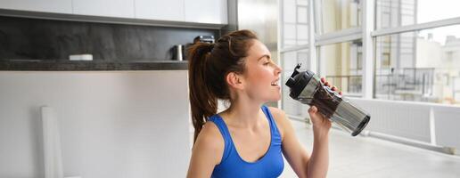 Portrait of sportswoman drinking water during workout session in living room, does her fitness training exercises at home photo