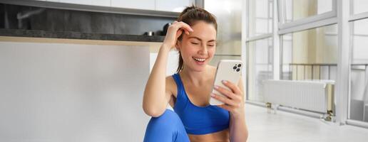 Image of young fit woman wearing sportbra and blue leggings, using smartphone app, does workout from home, sits on floor and follows online training video photo