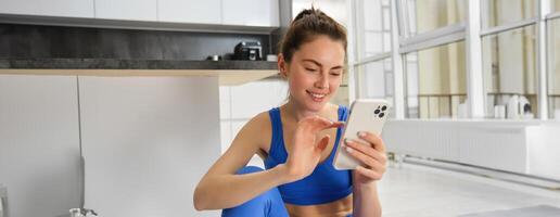 Young woman with smartphone does workout at home, using mobile phone app for sports training indoors, wears blue sportsbra and leggings photo