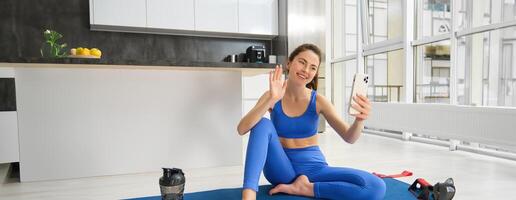 Portrait of young styling fitness girl doing workout from home, taking selfie and video for social media, gym instructor records her training session indoors photo