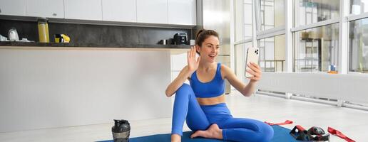 Image of beautiful, atheltic girl sits on yoga mat at home with smartphone, takes selfie and workout, does fitness exercises photo