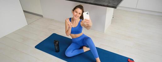 Portrait of stylish, smiling young brunette woman, wears blue sportswear, poses on yoga mat with smartphone, shows peace sign and takes selfie, does workout at home photo