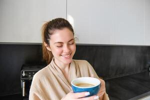 Lifestyle concept. Portrait of happy brunette woman in bathrobe, drinking coffee in the kitchen, having morning cuppa and smiling photo