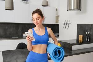 Portrait of attractive, fit young woman in sportswear, standing in living room with rubber yoga mat, holding smartphone, doing fitness exercises with mobile app at home photo