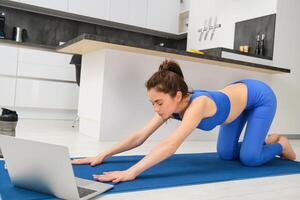 Portrait of woman doing stretching exercises, follows tutorial on laptop, attends online yoga class from home, workout on rubber mat photo