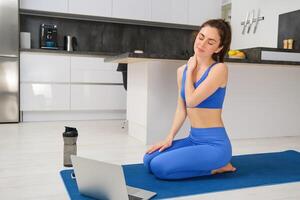 Portrait of fit and healthy woman doing fitness exercises, online yoga class from home, sitting with laptop on sports mat and massaging her neck photo