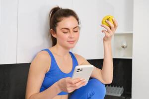 Portrait of brunette fitness woman, eating an apple, holding smartphone, using mobile phone app while having healthy fruit snack in kitchen photo