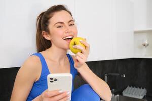 Portrait of smiling brunette girl, wearing fitness clothes, sitting in kitchen and biting an apple, eating fruit snack between workout, holding smartphone, using mobile phone app photo