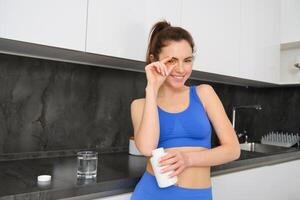 retrato de saludable, sonriente aptitud mujer, demostración vitaminas, pescado petróleo dietético suplemento para fuerte, ajuste cuerpo, Bebiendo agua con tableta, en pie en ktichen foto