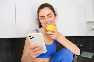 Portrait of sportswoman, girl eating and apple and looking at her social media, smartphone screen, having a snack in kitchen, wearing fitness activewear photo