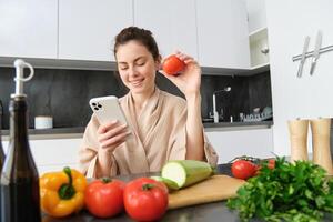 Young woman orders groceries on mobile app. Girl in bathrobe sits in the kitchen with vegetables, looking for recipe to cook dinner, using smartphone application photo